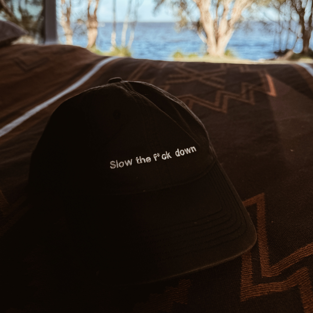 Black AS Colour cap with “Slow the Fck Down” embroidered on the front, photographed on a neutral background in natural light.
